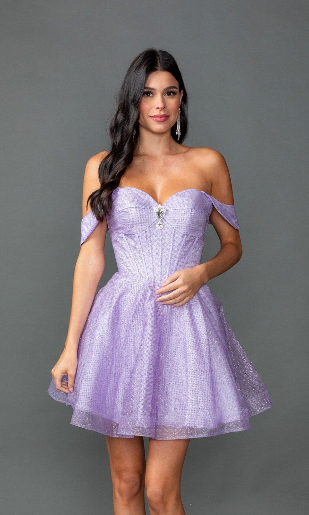 A woman poses in front of a plain gray background, wearing the Dancing Queen 3425—a short, off-the-shoulder lavender sparkly prom dress with a fitted bodice and flared skirt.